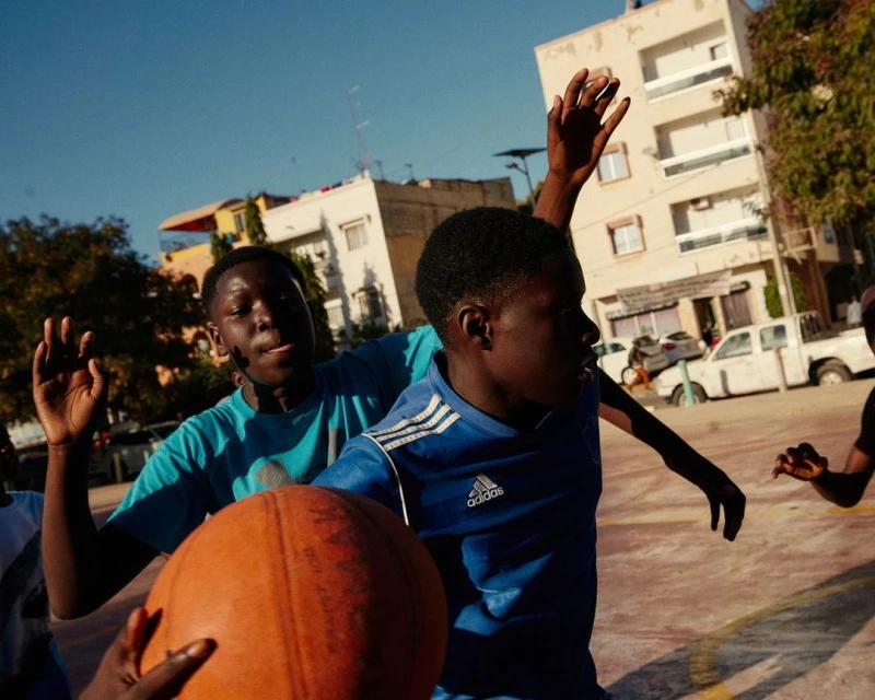Outdoor action shot of two young Black men playing basketball on a court in an urban setting during the daytime, with light blue sky.