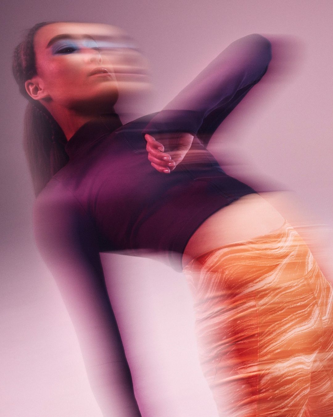 Abstract long exposure of a female fashion model in dark top and patterned skirt against a pink background in a studio setting.