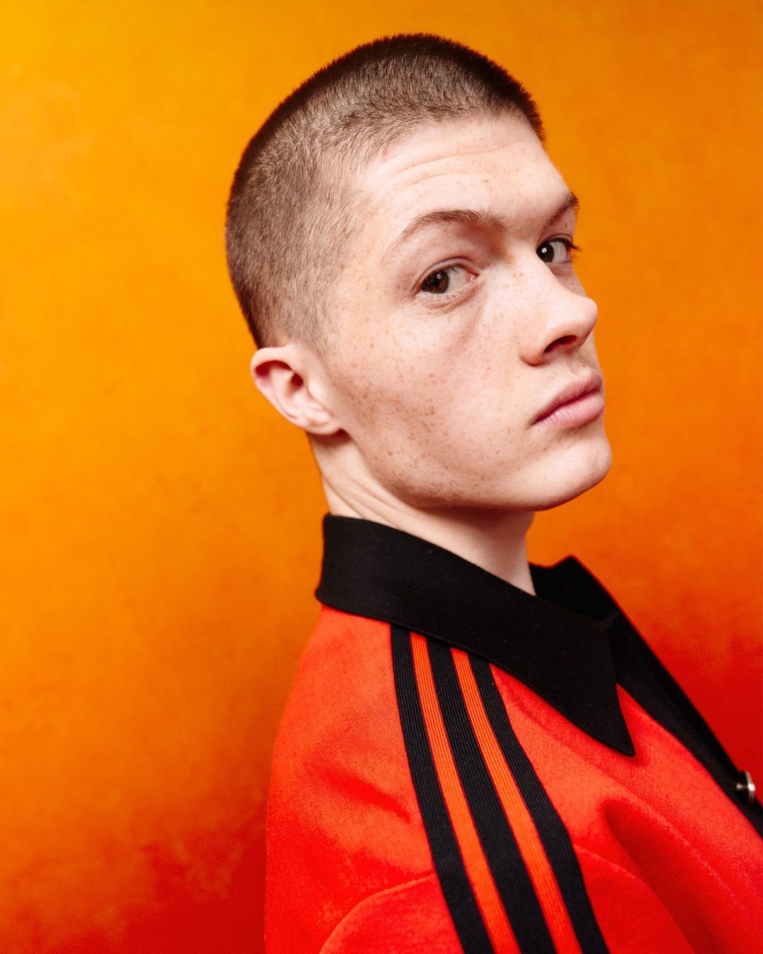 Portrait of a young adult male with a buzz cut wearing an orange jacket with black stripes, against a blended orange background.