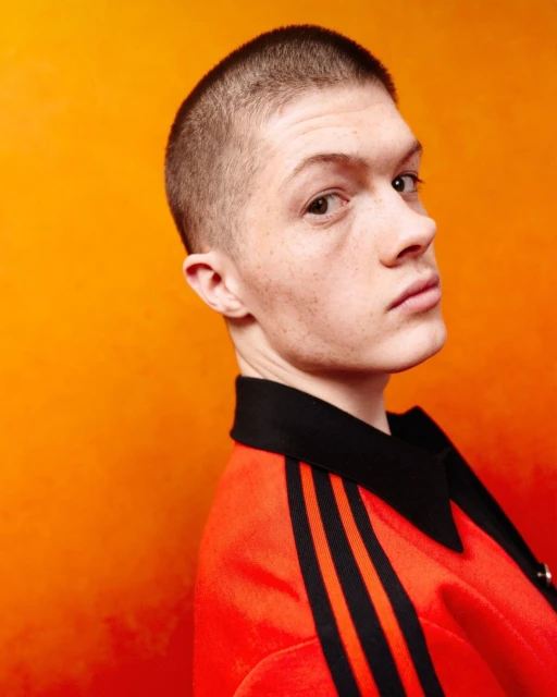 Portrait of a young adult male with a buzz cut wearing an orange jacket with black stripes, against a blended orange background.