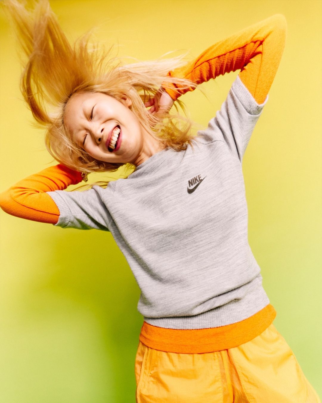 Smiling woman with blonde hair tossing her head back, wearing a grey Nike sweatshirt and orange pants in a photo studio setting.