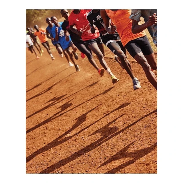 A group of runners compete outdoors on a dirt track with long shadows. The runners are wearing athletic gear and running shoes in bright daylight.