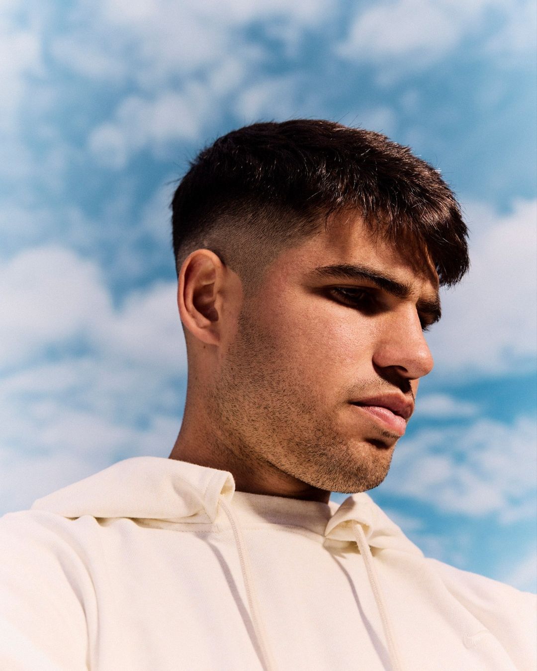 Portrait of handsome young man with dark hair and light skin wearing white hoodie, against a light blue cloudy sky background.