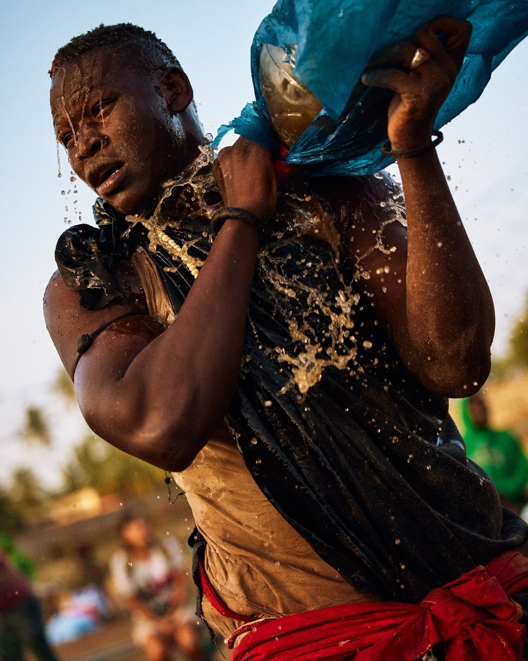 African man outdoors in dynamic action, pouring water from a blue plastic bag onto himself, captured in a medium shot with natural light.