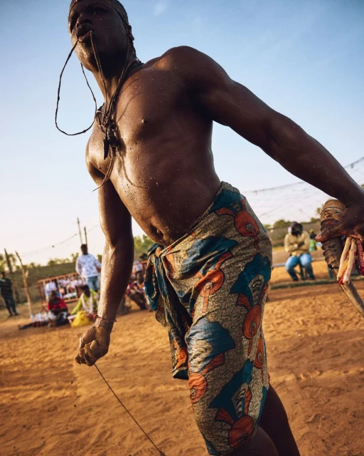 African man in traditional clothing performs ritual with rope and musical instrument in outdoor setting under blue sky in daytime.