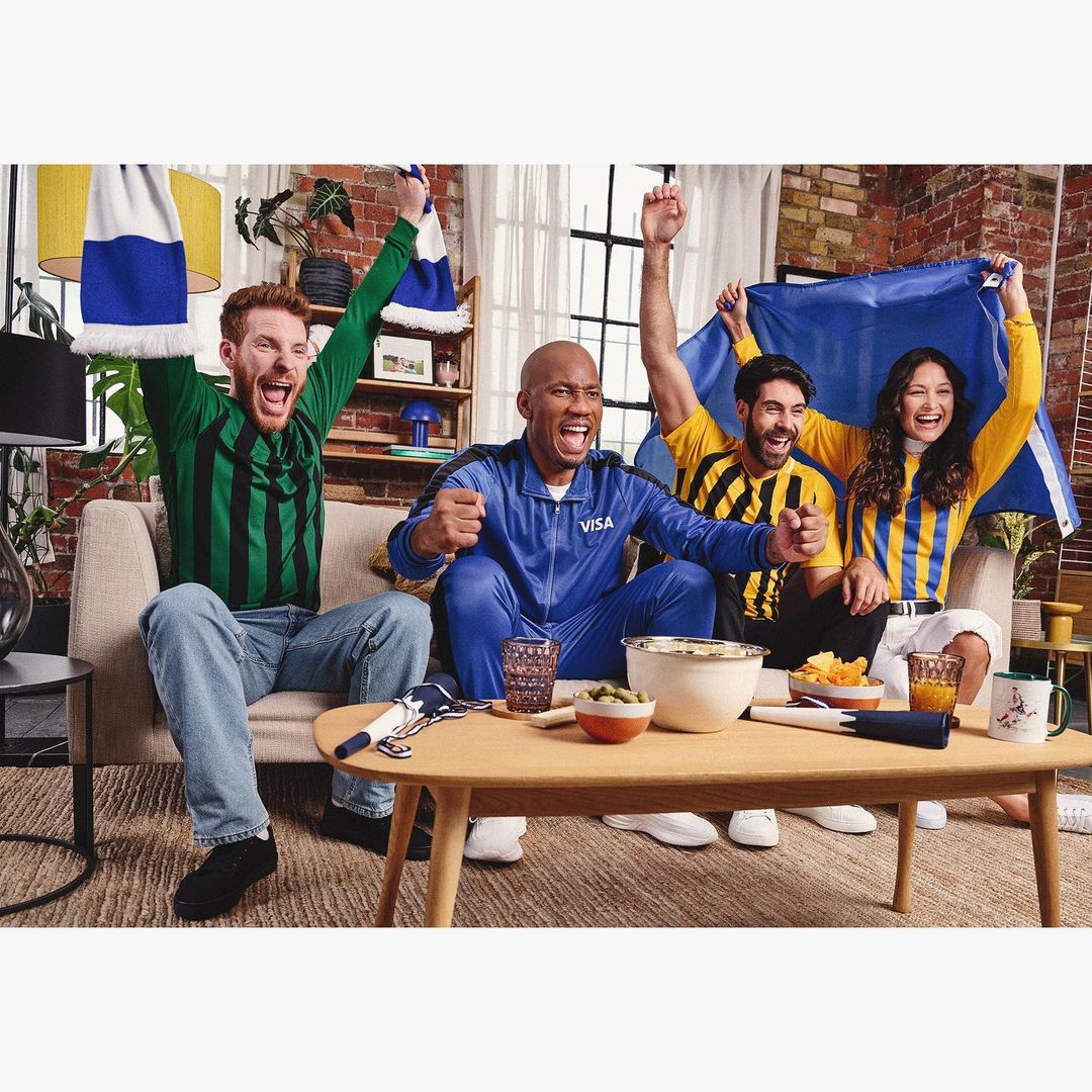 Five sports fans celebrate while watching a game in a living room, holding a scarf and a flag, with a table of snacks in front.