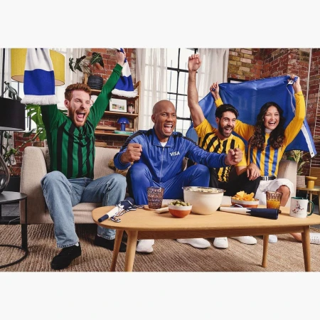 Five sports fans celebrate while watching a game in a living room, holding a scarf and a flag, with a table of snacks in front.