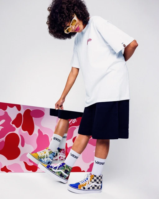 A stylish woman with curly hair is posing in a studio, wearing a white T-shirt, dark shorts, colorful high-top sneakers, and yellow sunglasses.