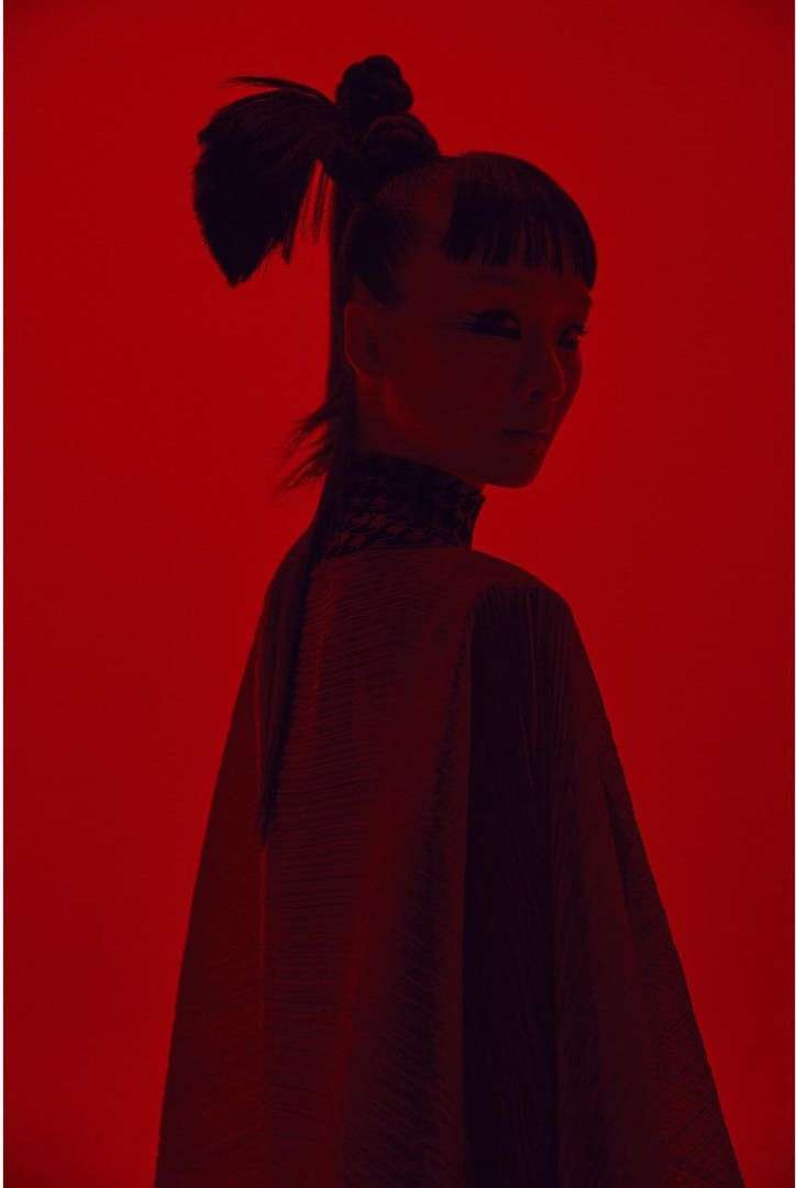 Portrait of a woman with a ponytail and decorative collar against a solid red background in a studio environment with dramatic lighting.