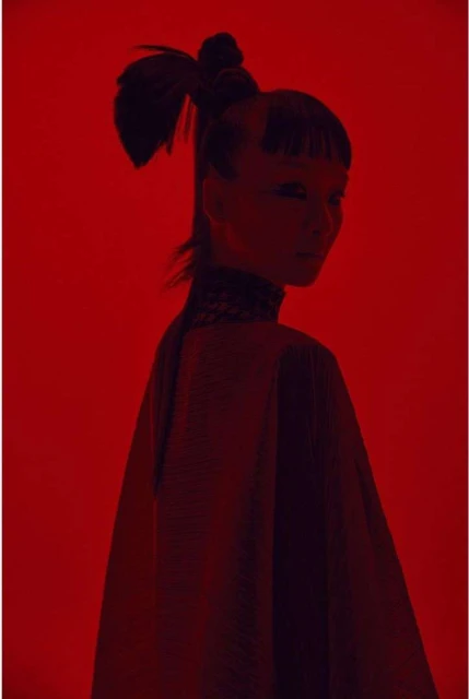 Portrait of a woman with a ponytail and decorative collar against a solid red background in a studio environment with dramatic lighting.