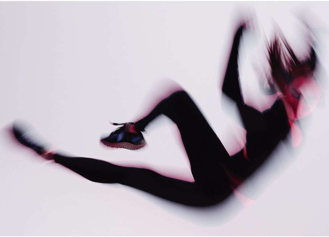 Motion blur photograph of a woman in black leggings and sneakers in a dynamic pose against a white background.