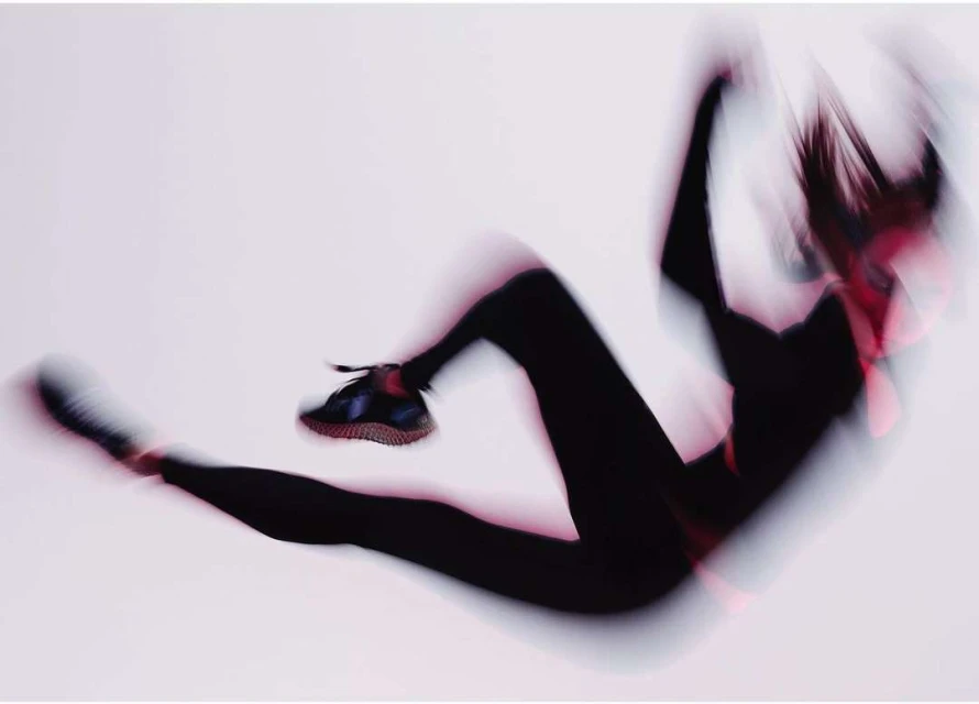 Motion blur photograph of a woman in black leggings and sneakers in a dynamic pose against a white background.