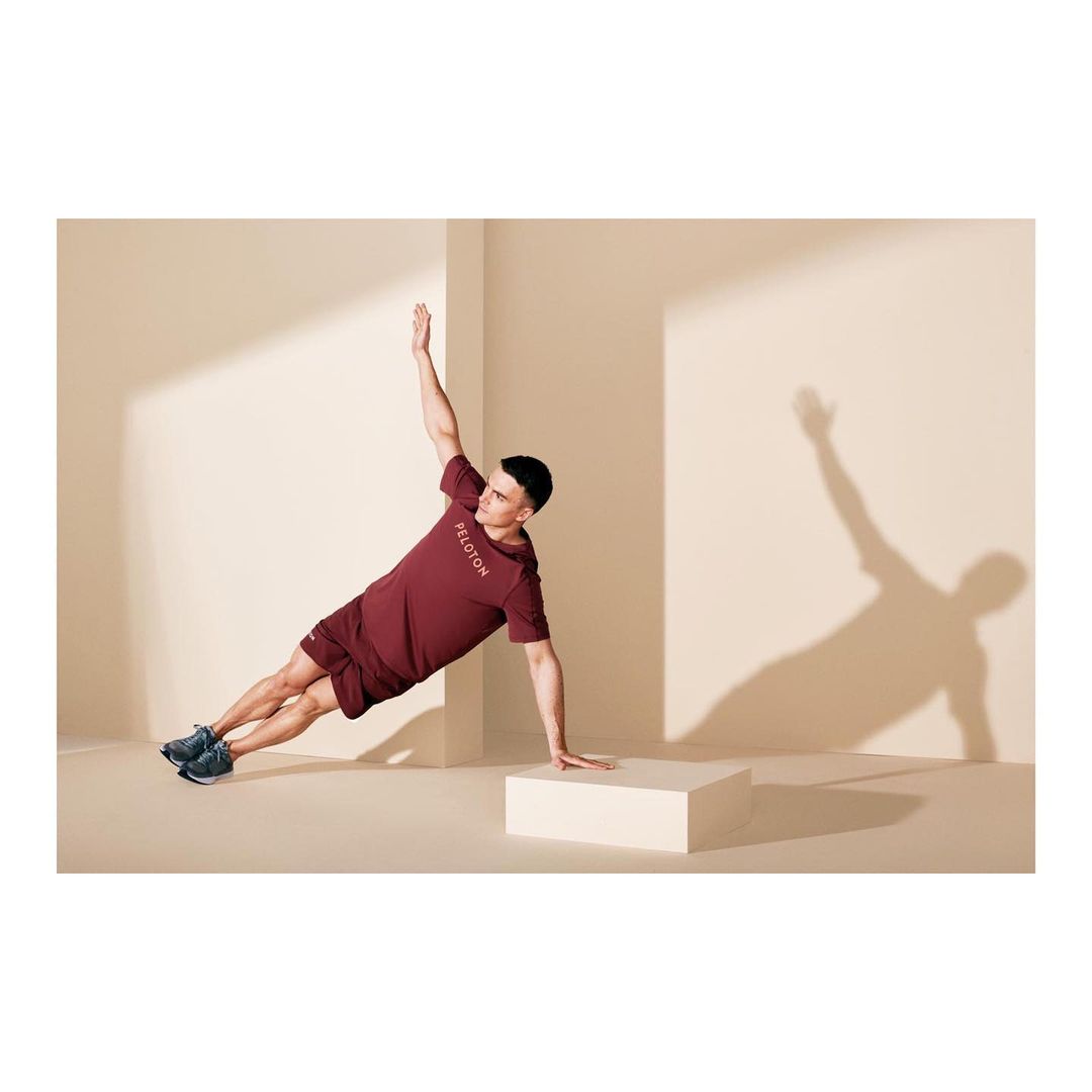 Athletic man in burgundy activewear performs side plank on a white block in a photo studio with neutral walls and natural light.