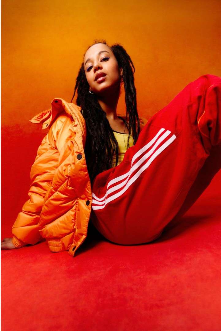 Portrait of a young woman with braided hair wearing an orange jacket and red Adidas pants, posing against an orange background.