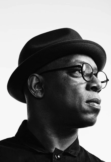 Black and white portrait of an African American man wearing a fedora and round glasses against a bright white background.