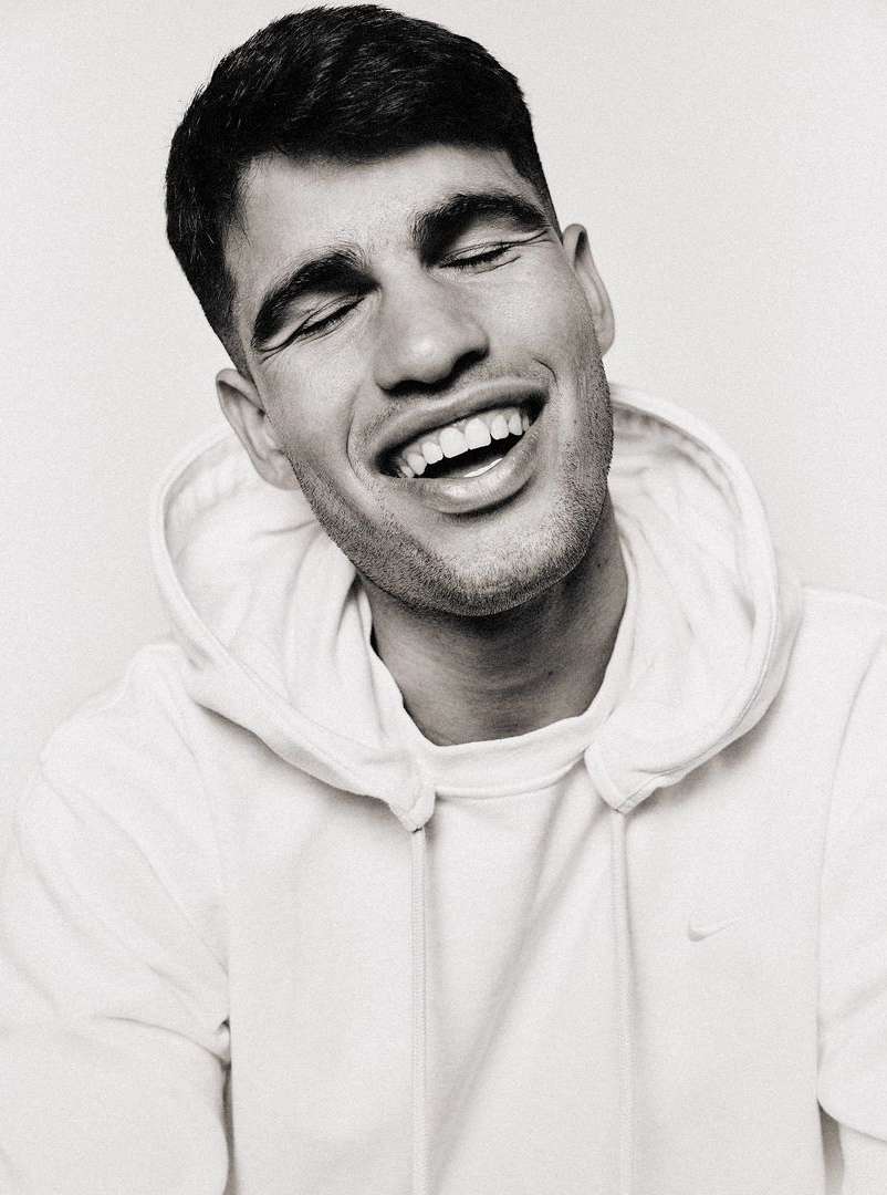 Close-up black and white portrait of a smiling adult man with closed eyes, wearing a hooded sweatshirt in an indoor setting.