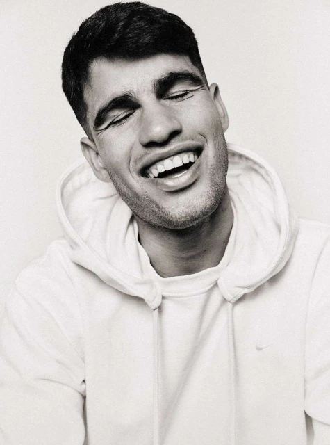 Close-up black and white portrait of a smiling adult man with closed eyes, wearing a hooded sweatshirt in an indoor setting.