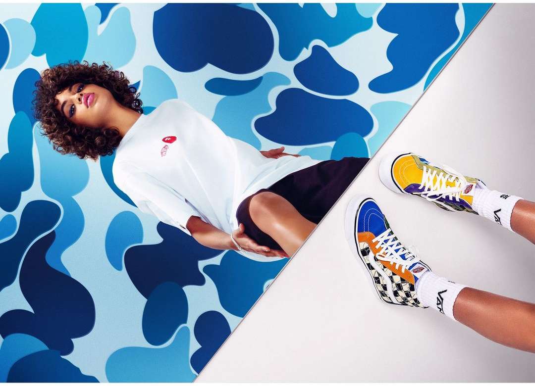 Eye-level shot of a woman lying on a split blue camo/white background wearing a white t-shirt, black shorts, and colorful hi-top sneakers.