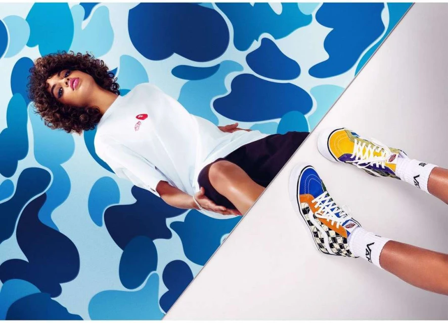 Eye-level shot of a woman lying on a split blue camo/white background wearing a white t-shirt, black shorts, and colorful hi-top sneakers.