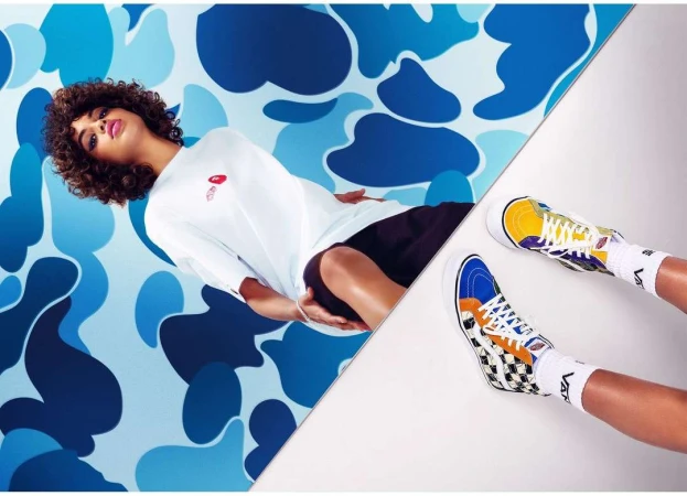Eye-level shot of a woman lying on a split blue camo/white background wearing a white t-shirt, black shorts, and colorful hi-top sneakers.