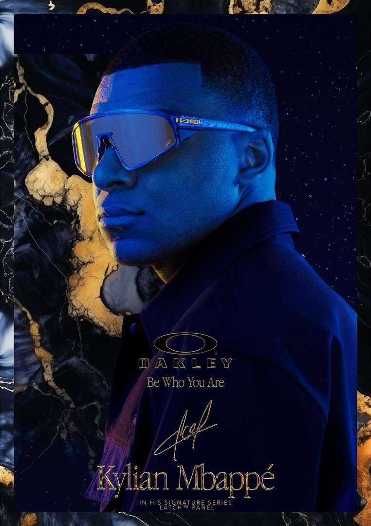 Close up studio shot of Kylian Mbappé wearing reflective blue glasses and a blue jacket against a dark marble background.