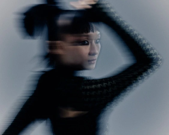 Motion blurred portrait of an East Asian woman wearing a patterned long sleeve top with dark eye makeup against a grey background.