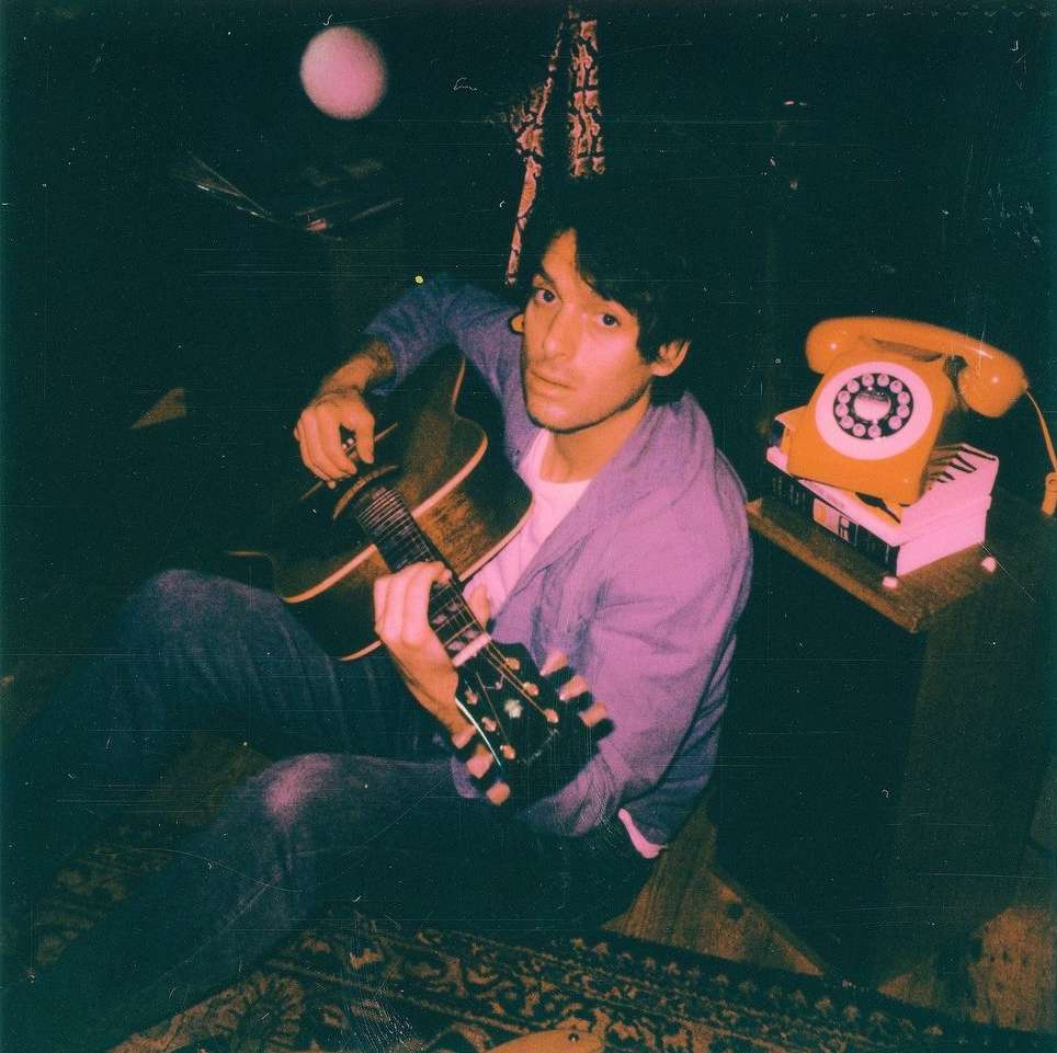 A man sits indoors playing an acoustic guitar, illuminated by soft, pinkish lighting, with a rotary telephone and books nearby.