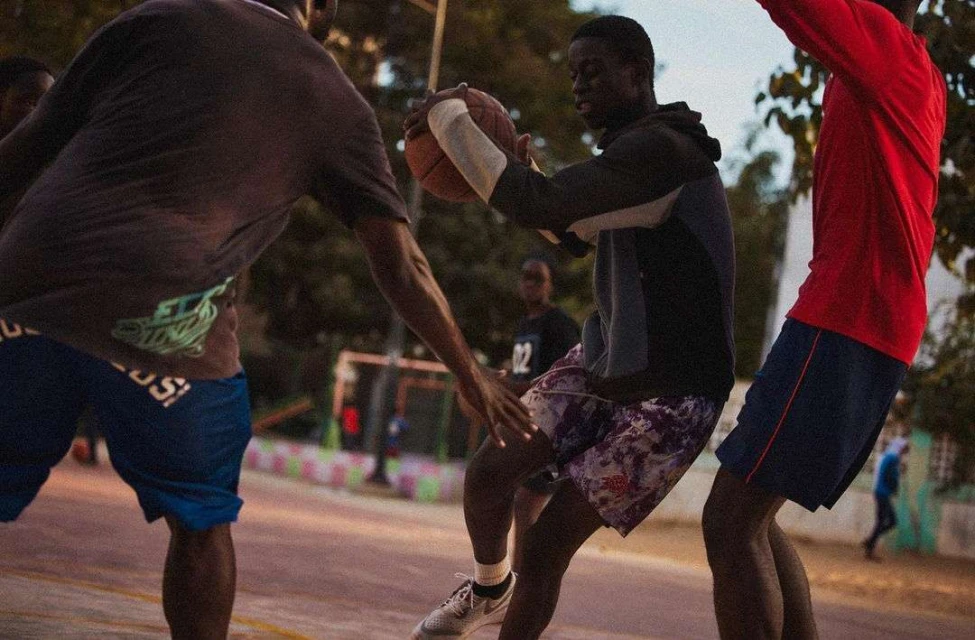 Group of young adult males playing basketball outdoors on a court, evening light, athletic wear, casual setting.