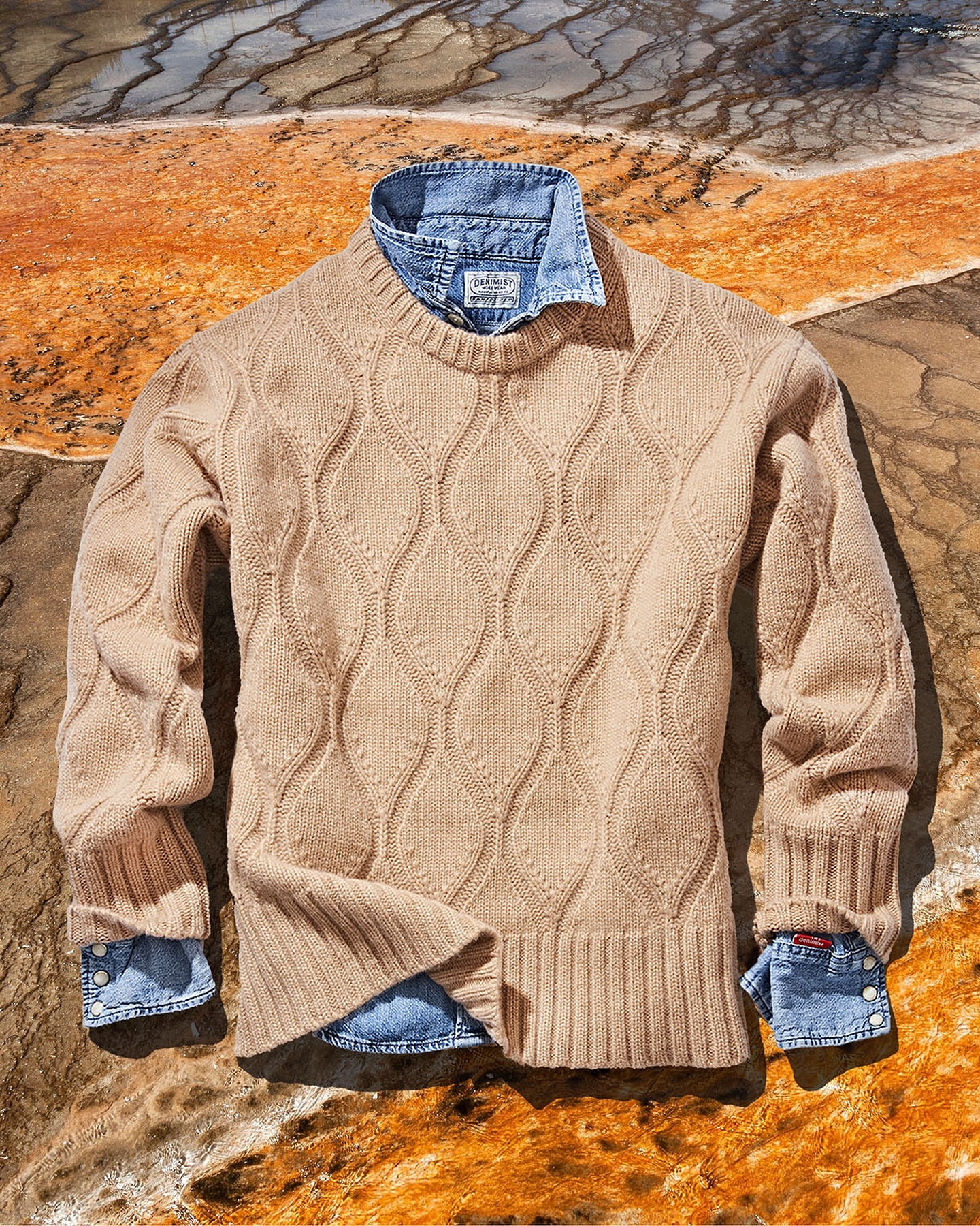 Tan knit sweater layered over a blue denim shirt on a textured stone background, showcasing neutral tones and outdoor fashion.