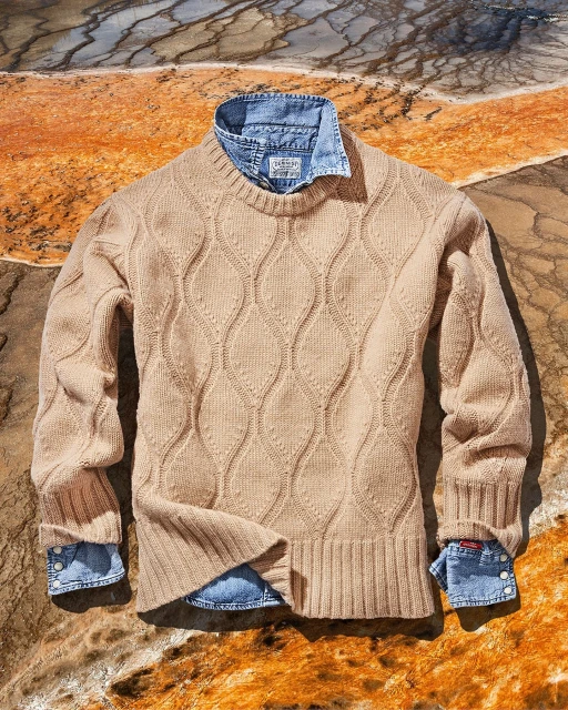 Tan knit sweater layered over a blue denim shirt on a textured stone background, showcasing neutral tones and outdoor fashion.