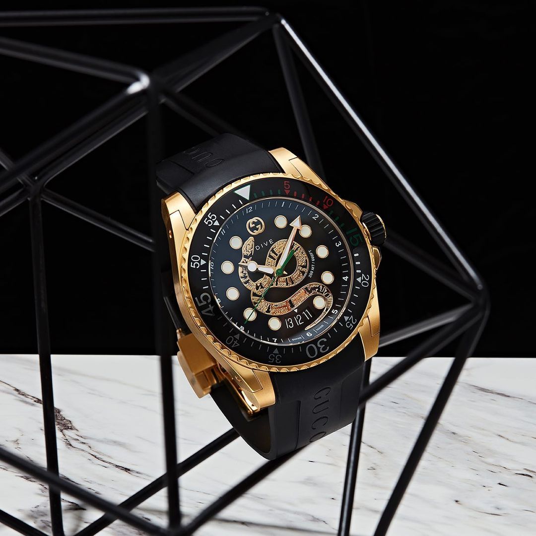 Gold Gucci Dive watch with black band and snake motif on a marble surface, presented in a graphic black wireframe for a modern look.