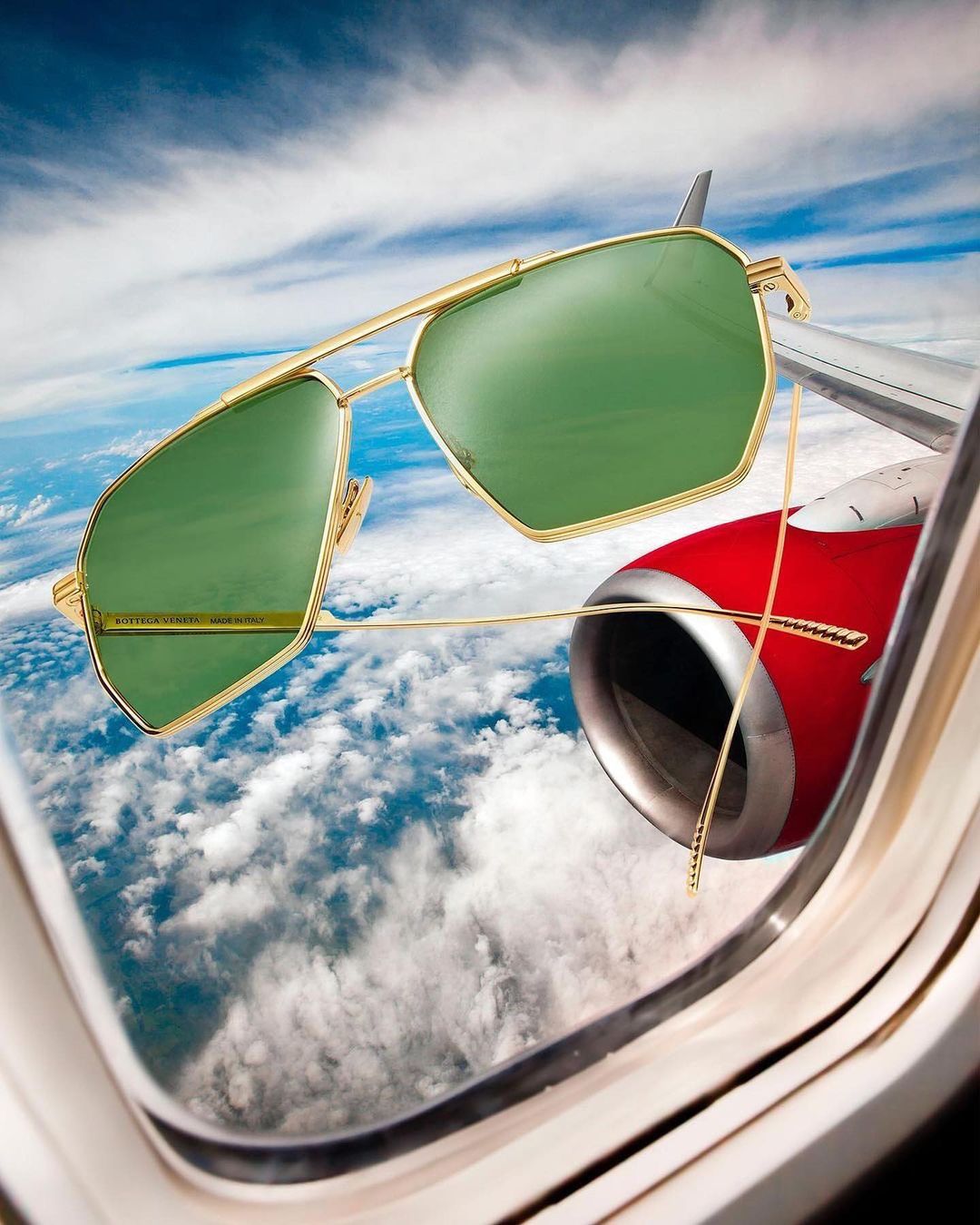 Gold sunglasses with green lenses are positioned against the backdrop of an airplane wing, engine, and cloudy sky visible through the window.