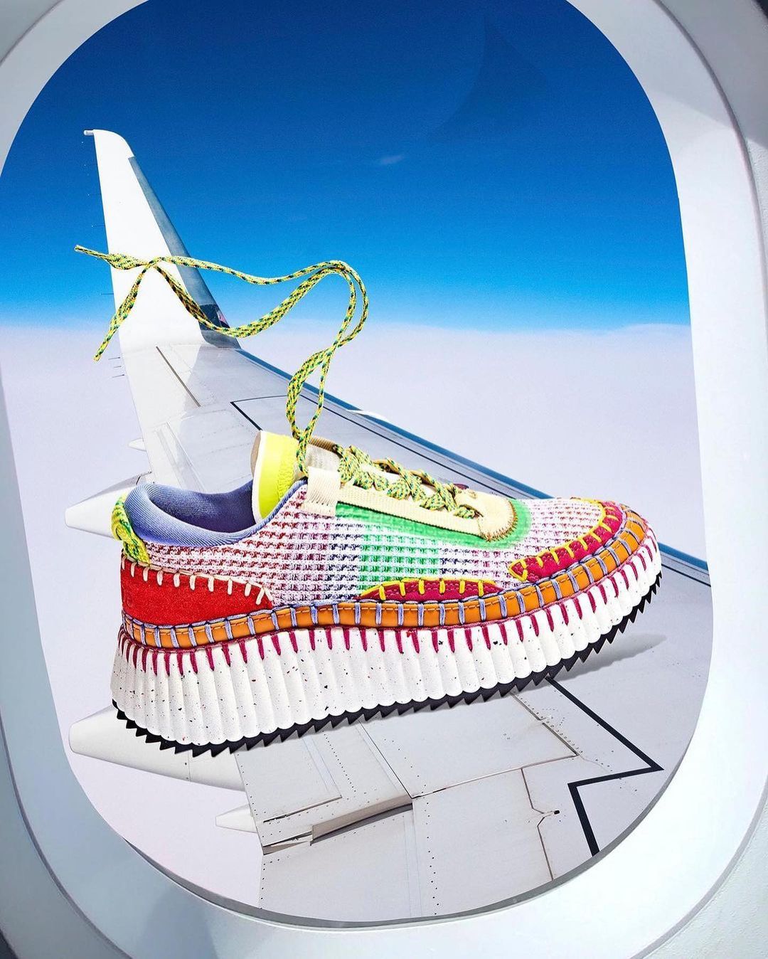 Elevated view of colorful sneaker displayed on airplane wing near window, against blue sky. Bright, modern product shot.