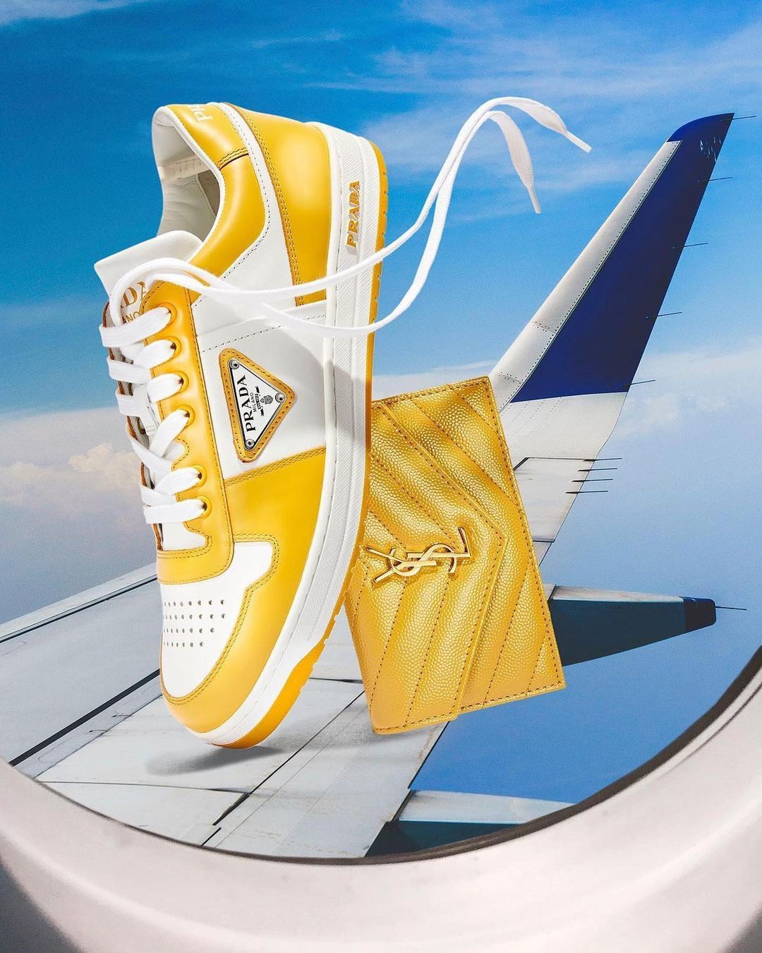 Elevated view of a yellow Prada sneaker and a yellow YSL wallet floating in front of an airplane wing against a bright blue sky.