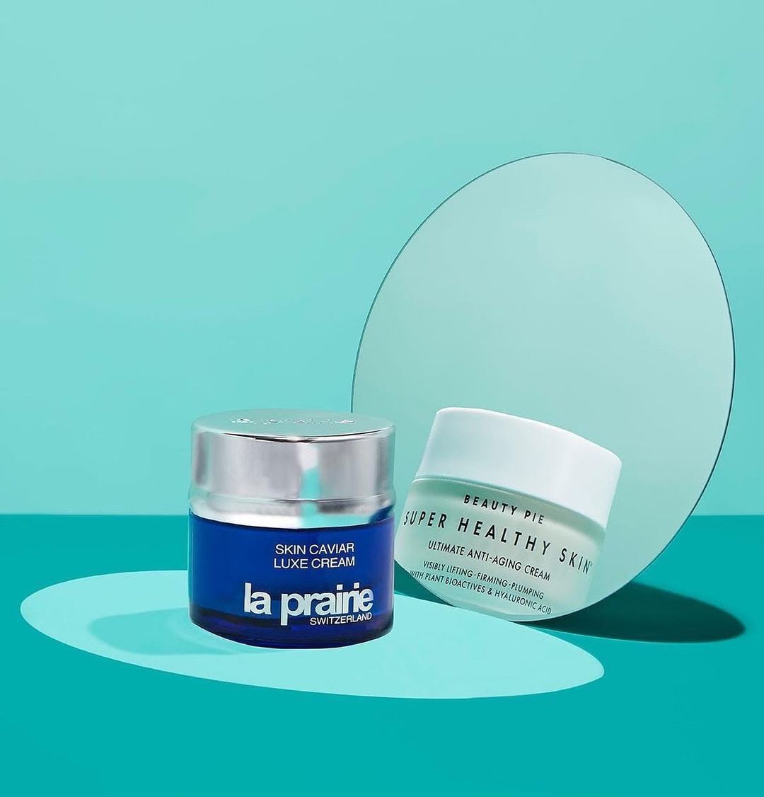 La Prairie and Beauty Pie anti-aging skin cream products, displayed against a teal background with a round mirror. Beauty products.