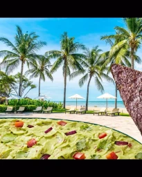 Close-up of a blue corn tortilla chip dipped into a bowl of chunky guacamole with a tropical beach and palm trees in the background.
