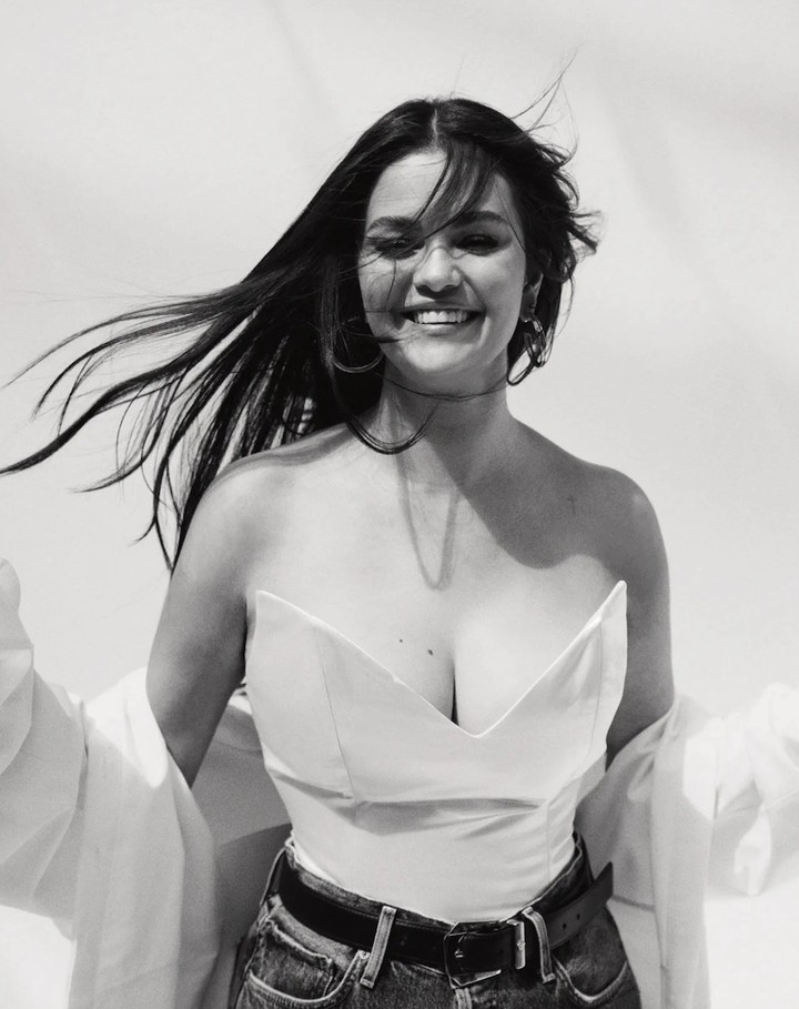 Portrait of Selena Gomez smiling with dark flowing hair in the wind wearing a white strapless top and denim jeans