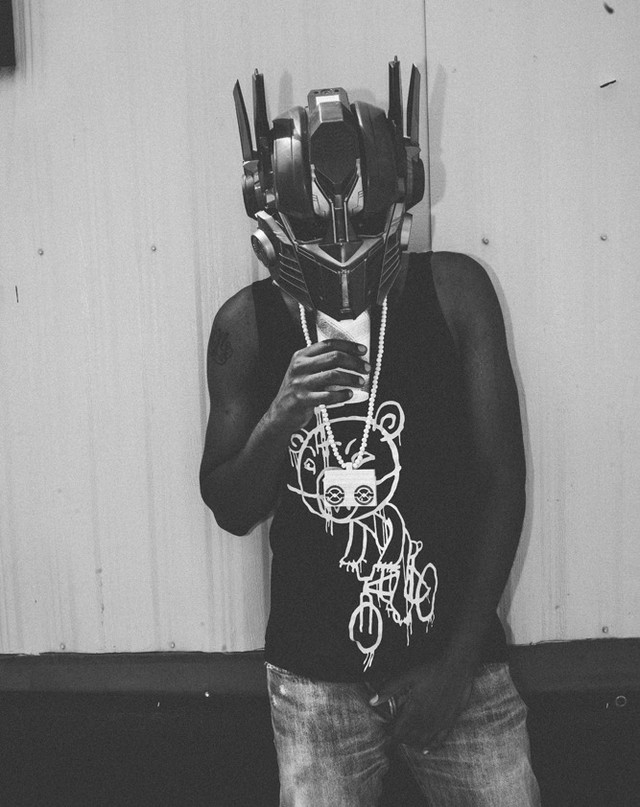 Adult male in tank top and jeans wearing a Transformers Optimus Prime mask standing indoors in black and white photo.