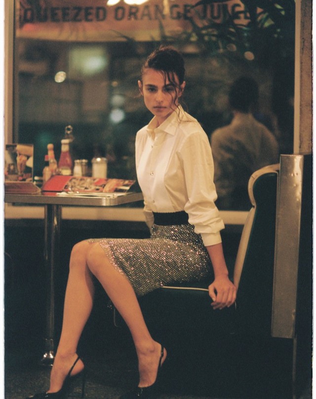 Fashionable woman poses in retro diner wearing white blouse and sparkling skirt. Black high heels complete her elegant look.