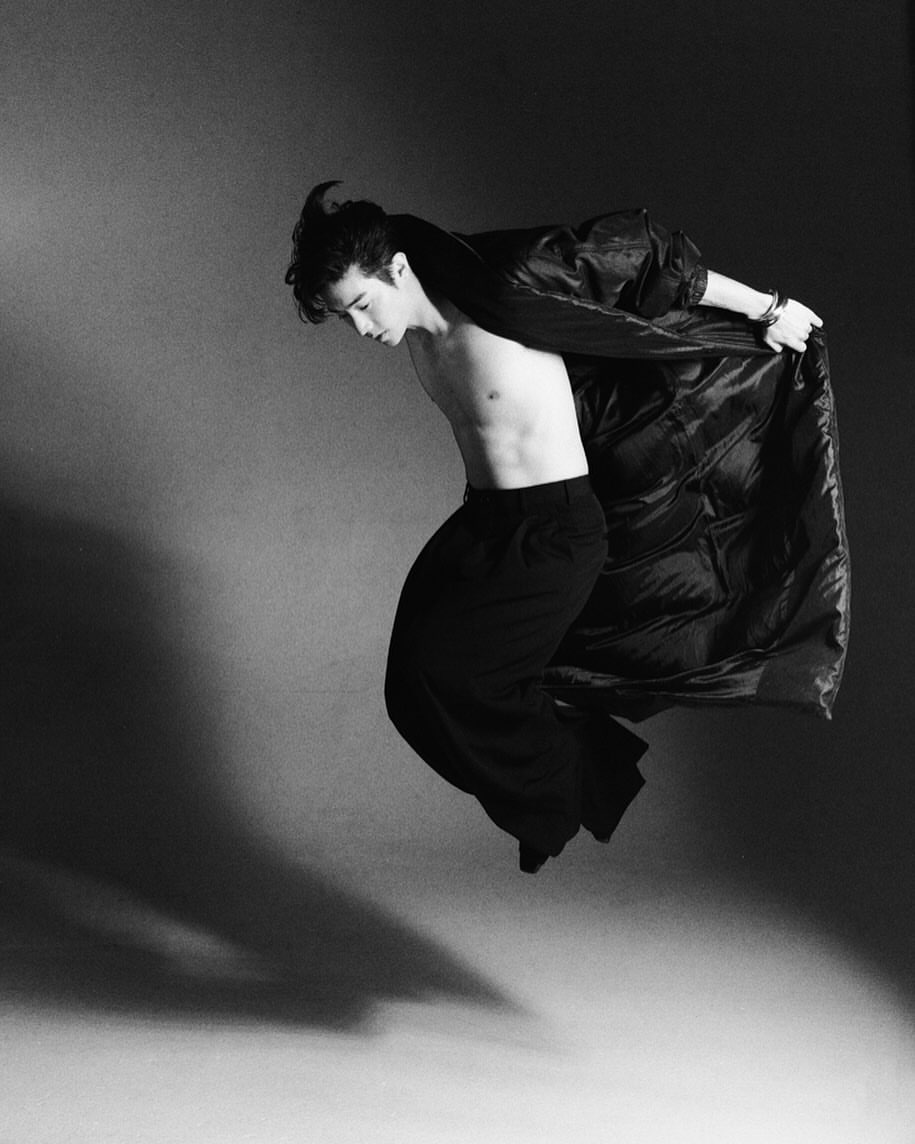 Monochrome portrait of a young adult male in mid-air, wearing black pants and a dramatic coat, against a dark grey studio backdrop with shadows.
