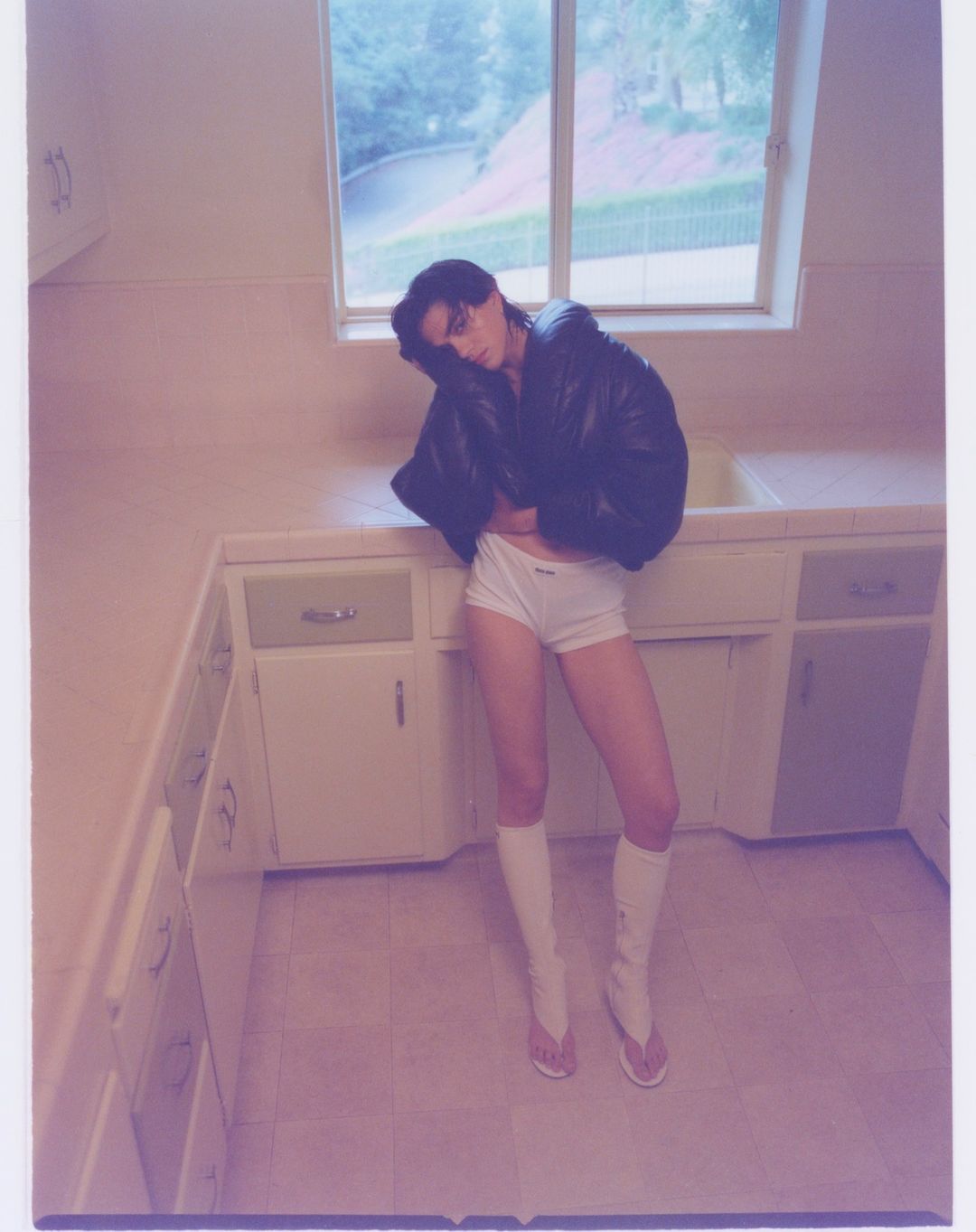 Woman in black jacket and white shorts posing in kitchen. Natural light from window, casual indoor fashion portrait, neutral color palette.