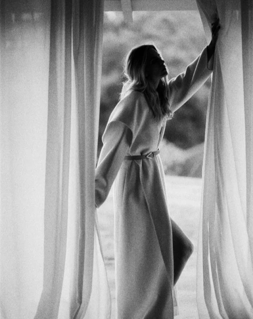 Black and white shot of a woman standing between sheer curtains in a flowing coat, looking upwards into the light from the outdoor scene.