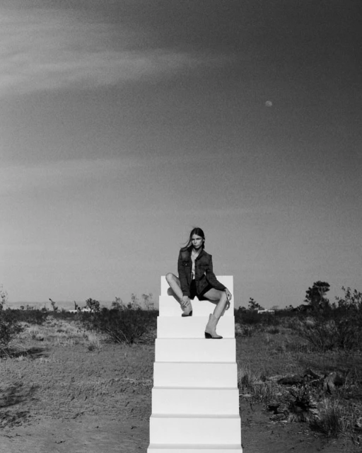 Black and white image of a woman in a jacket and shorts sitting on white stairs in a desert landscape under a bright sky.