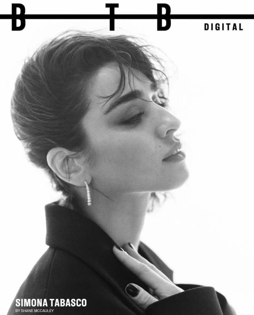 Black and white close-up portrait of a woman with short hair wearing a dark coat and diamond earring, looking upwards in a studio.