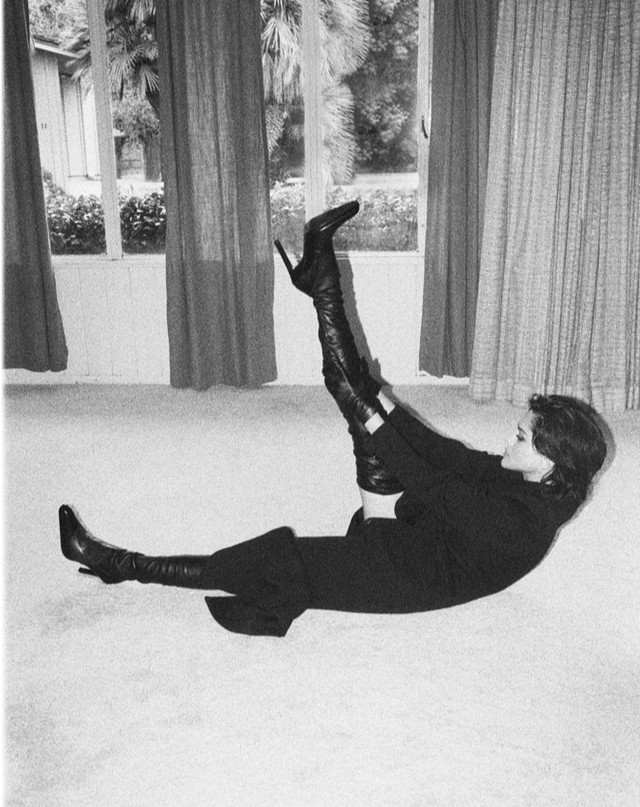 A woman in a dark outfit and thigh-high heeled boots is posed on the floor in front of a window with sheer curtains.