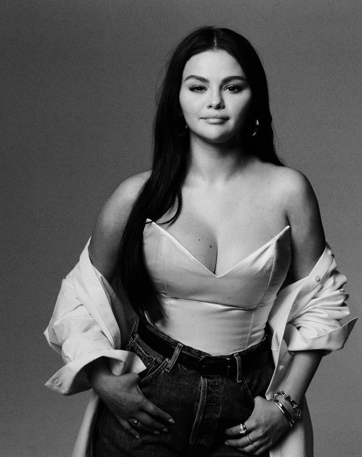 Monochrome studio portrait of Selena Gomez, wearing a bustier, jeans, and an open shirt, against a plain gray backdrop.