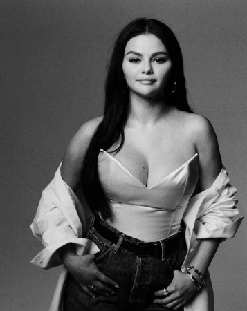 Monochrome studio portrait of Selena Gomez, wearing a bustier, jeans, and an open shirt, against a plain gray backdrop.
