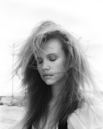 Monochrome portrait of a woman with voluminous, wind-swept hair and closed eyes against an outdoor background.