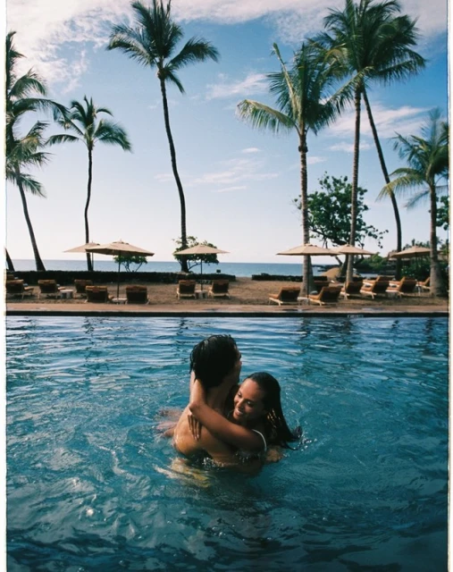 Couple embraces in a pool with turquoise water, with palm trees and beach umbrellas dotting the shoreline and clear blue sky.