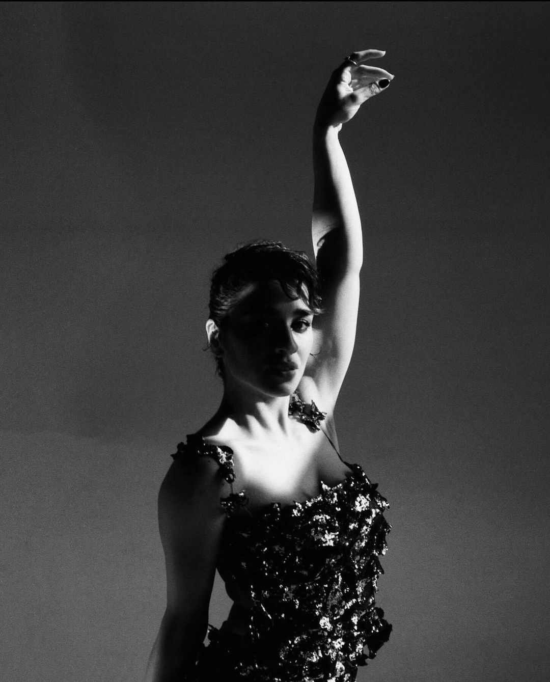 Monochrome portrait of a woman in a textured dress raising her arm in a dramatic pose against a gradient grey backdrop.